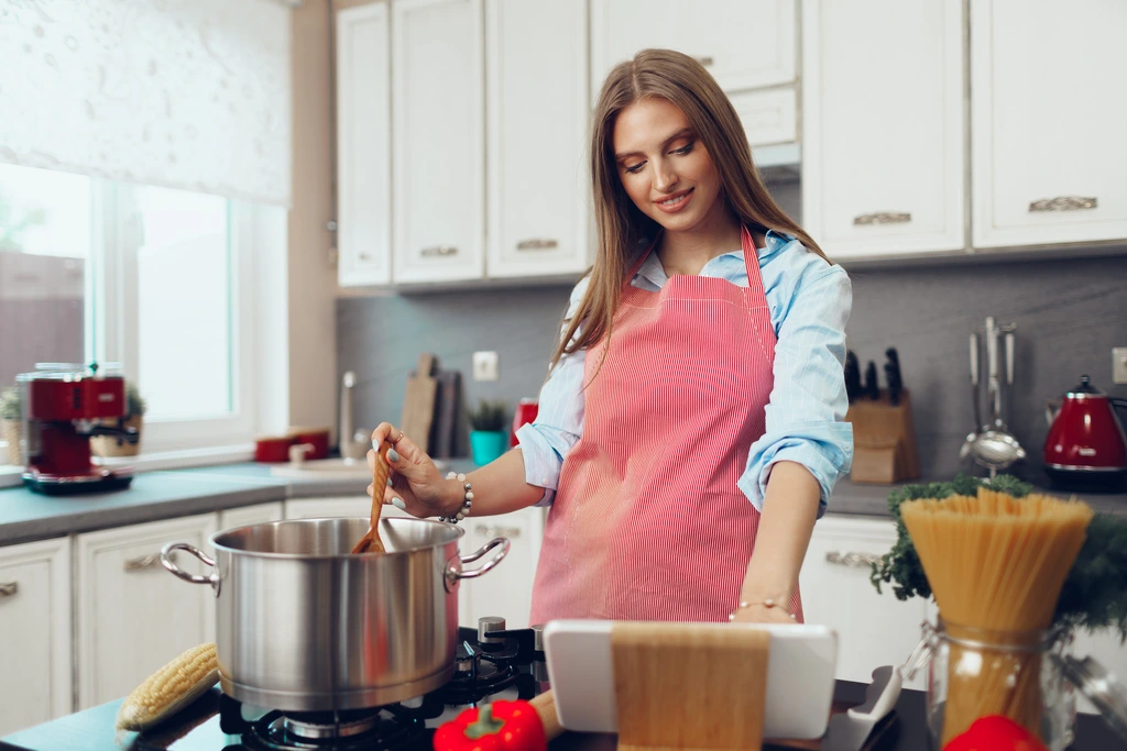 Amy Rosewood, author of Daisy's Recipes, smiles as she stirs a pot on the stove while looking at a recipe on a tablet in her bright, modern kitchen.