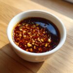 A small white bowl filled with rich, dark Dumpling Sauce topped generously with minced garlic and chili flakes.