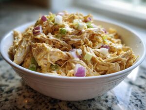 Close-up of shredded chicken mixed in a creamy dressing, topped with red onion and green onion, representing honey mustard chicken salad.