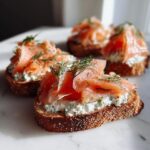 Close-up of an appetizer featuring smoked salmon piled high on cream cheese spread over toasted bread, garnished with fresh dill.