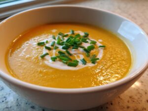 A close-up of a vibrant orange bowl of creamy cheese soup, topped with a swirl of cream and fresh chopped chives.