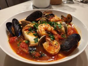 A close-up of a vibrant seafood soup featuring mussels, shrimp, clams, and a white fish fillet in a rich tomato broth.