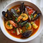 A close-up of a vibrant bowl of seafood soup featuring mussels, shrimp, white fish, and clams in a tomato broth.