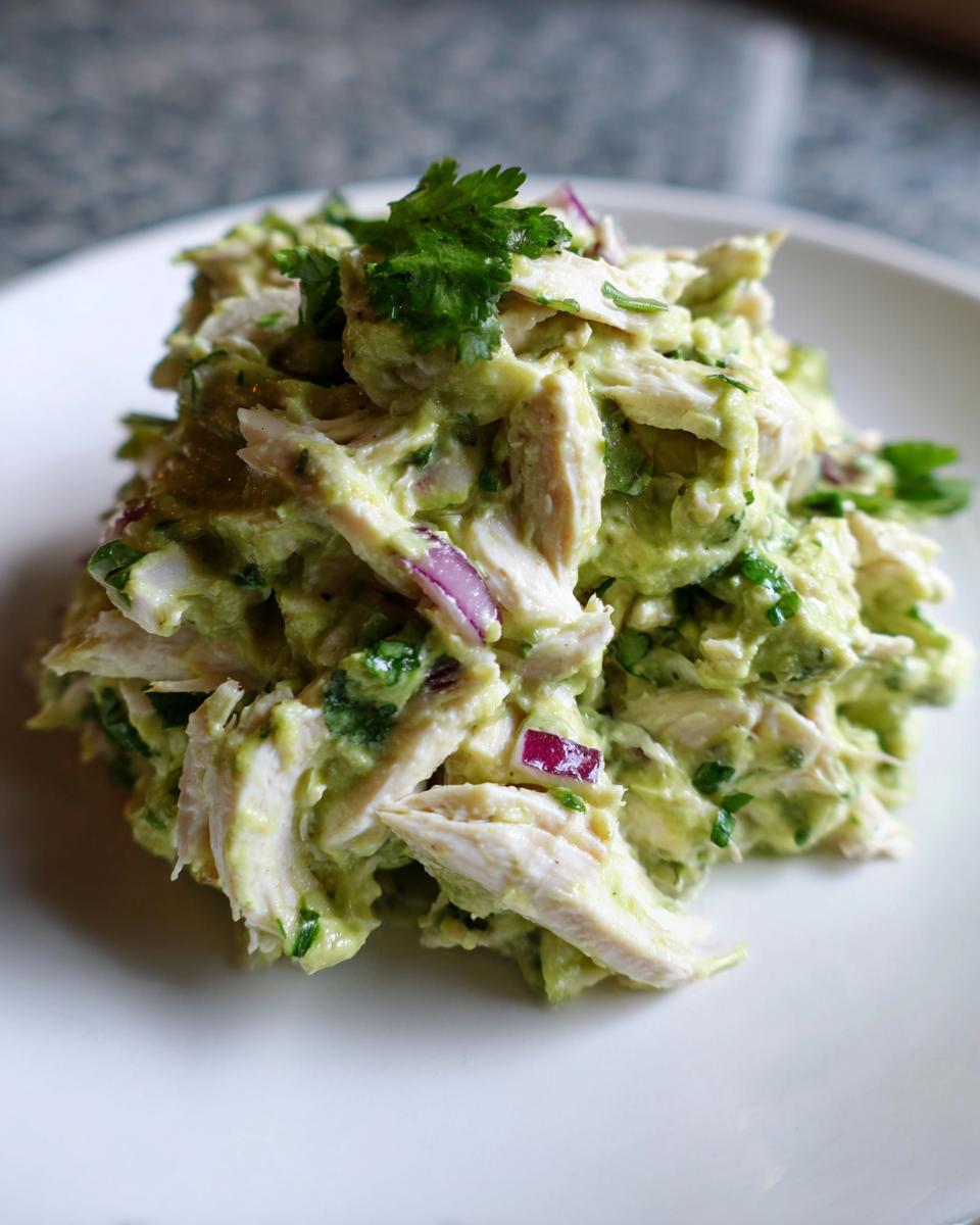 A mound of creamy avocado chicken salad mixed with shredded chicken, red onion, and cilantro on a white plate.