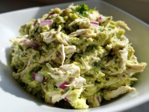 Close-up of creamy avocado chicken salad mixed with shredded chicken and diced red onion in a white bowl.
