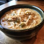 Close-up of a dark bowl filled with creamy, shredded cream cheese chicken chili featuring white beans and green peppers.