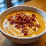 Close-up of a bowl of creamy orange cheeseburger soup recipe garnished with crispy bacon bits and a swirl of cream.