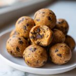 A stack of homemade chocolate chip protein balls on a white plate, with one ball bitten to show the texture.