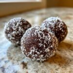 Three rich chocolate rum balls rolled in shredded white coconut flakes resting on a granite countertop.
