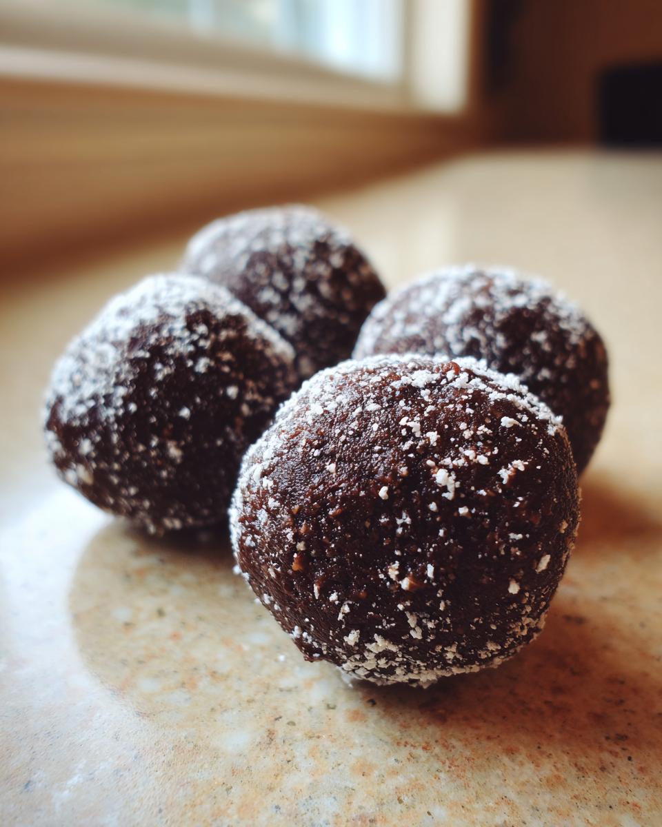 Four dark chocolate rum balls dusted with white powdered sugar resting on a light countertop.