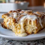 A close-up of a moist slice of cinnamon roll casserole covered in thick, dripping vanilla icing on a white plate.