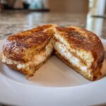 A golden brown, thick slice of cream cheese stuffed french toast cut in half, revealing the melted white filling.
