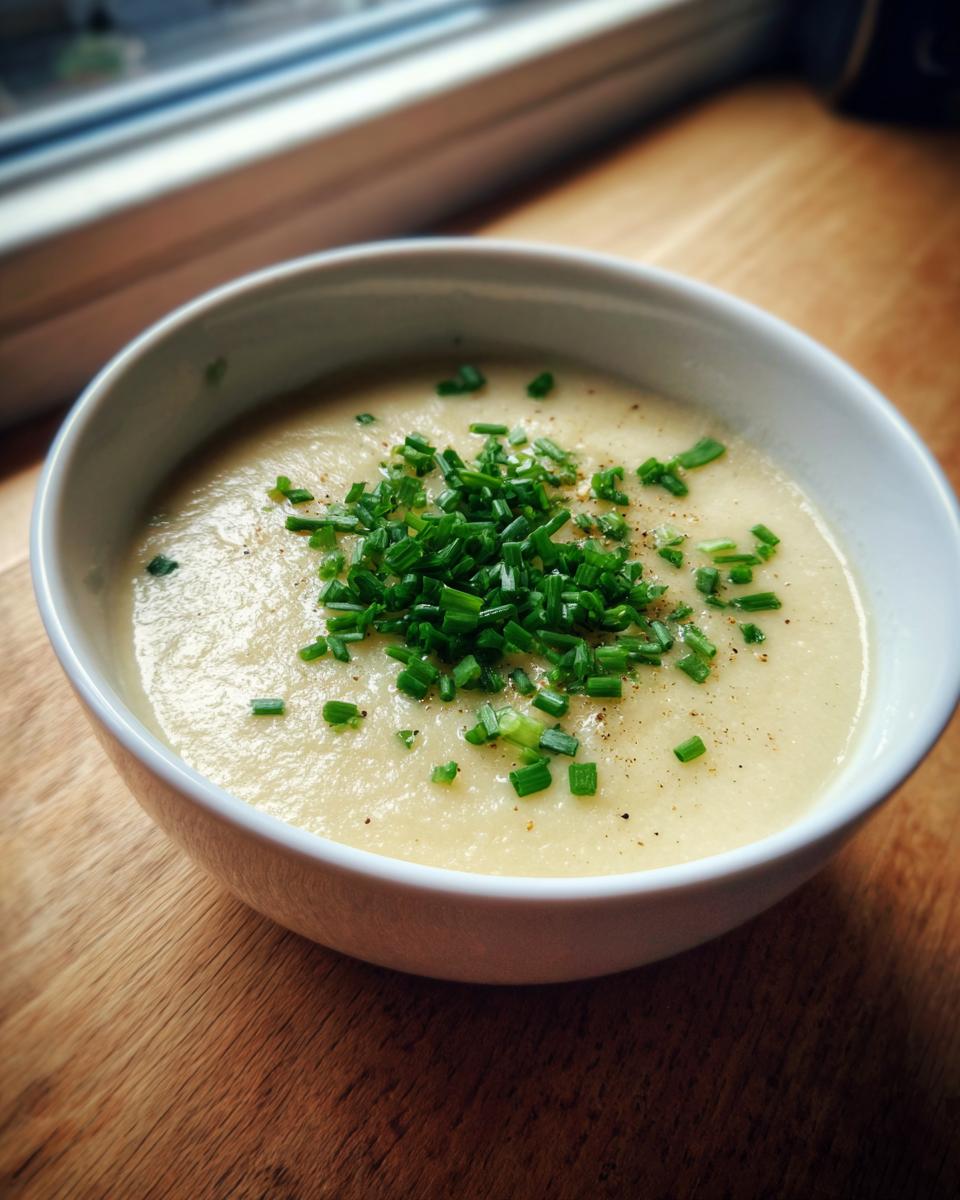 A close-up of creamy cauliflower soup in a white bowl, garnished generously with chopped green chives and black pepper.