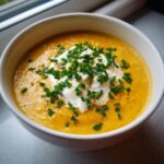 Close-up of a vibrant orange cheese soup topped with a dollop of sour cream and fresh chopped chives.