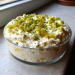 A creamy, white dirty martini dip topped with chopped green olives and fresh herbs in a clear glass bowl.
