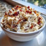 Close-up of a white bowl filled with creamy Neiman Marcus dip topped generously with crispy bacon pieces.
