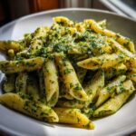 A close-up, appetizing shot of penne pasta coated in rich, creamy pesto pasta sauce in a white bowl.