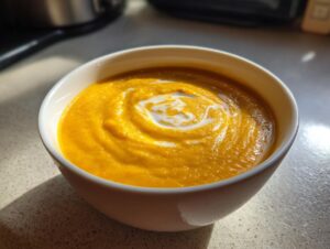 A close-up of a white bowl filled with vibrant orange pumpkin soup, swirled with cream.