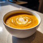 A close-up of vibrant orange pumpkin soup garnished with a swirl of cream and cracked black pepper.
