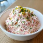 A close-up of creamy pink salmon dip topped with fresh dill and bright yellow lemon zest in a white bowl.