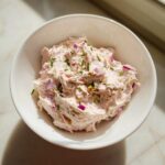 A white bowl filled with creamy, pink tuna dip, topped with chopped red onion and fresh chives.