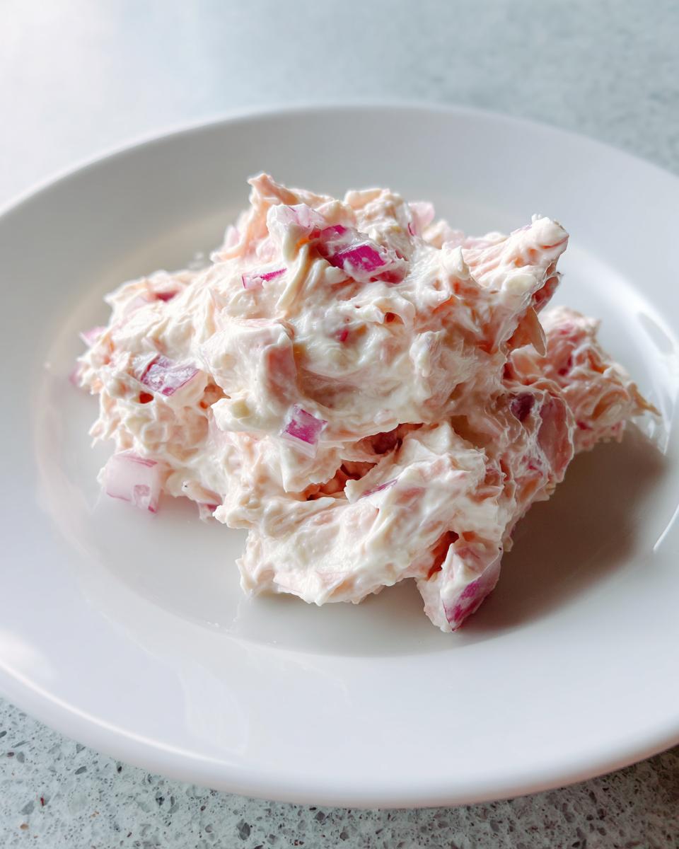 A creamy mound of pinkish tuna dip mixed with visible chunks of bright red onion served on a white plate.