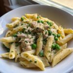 Close-up of creamy tuna pasta featuring penne coated in sauce, topped with shredded tuna, bright green peas, and fresh parsley.