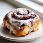 A close-up of a single, freshly baked cinnamon roll topped with thick white icing, sitting on a small plate.
