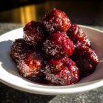 A close-up of several glossy cranberry meatballs piled high in a white bowl, coated in a rich, dark red sauce.