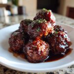 A close-up of several glistening Grape Jelly Meatballs piled on a white plate, covered in a rich, dark glaze and sprinkled with parsley.