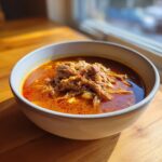 A close-up of a hearty bowl of Pozole rojo topped generously with shredded pork, sitting on a wooden table near a bright window.