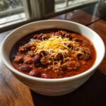 Close-up of a white bowl filled with rich, dark red crockpot chili topped with shredded cheddar and Monterey Jack cheese.