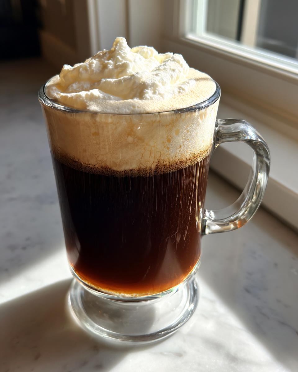 A tall glass mug filled with dark coffee, topped with a thick layer of cream and whipped cream, characteristic of an Irish Coffee.