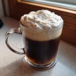A rich, dark Irish Coffee topped generously with fluffy whipped cream in a clear glass mug.