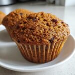 Close-up of a perfectly baked, moist bran muffins with visible raisins, sitting on a white plate.