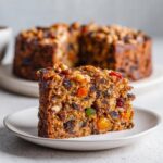 A close-up of a moist slice of fruit cake, packed with dried and candied fruits and nuts, served on a white plate.