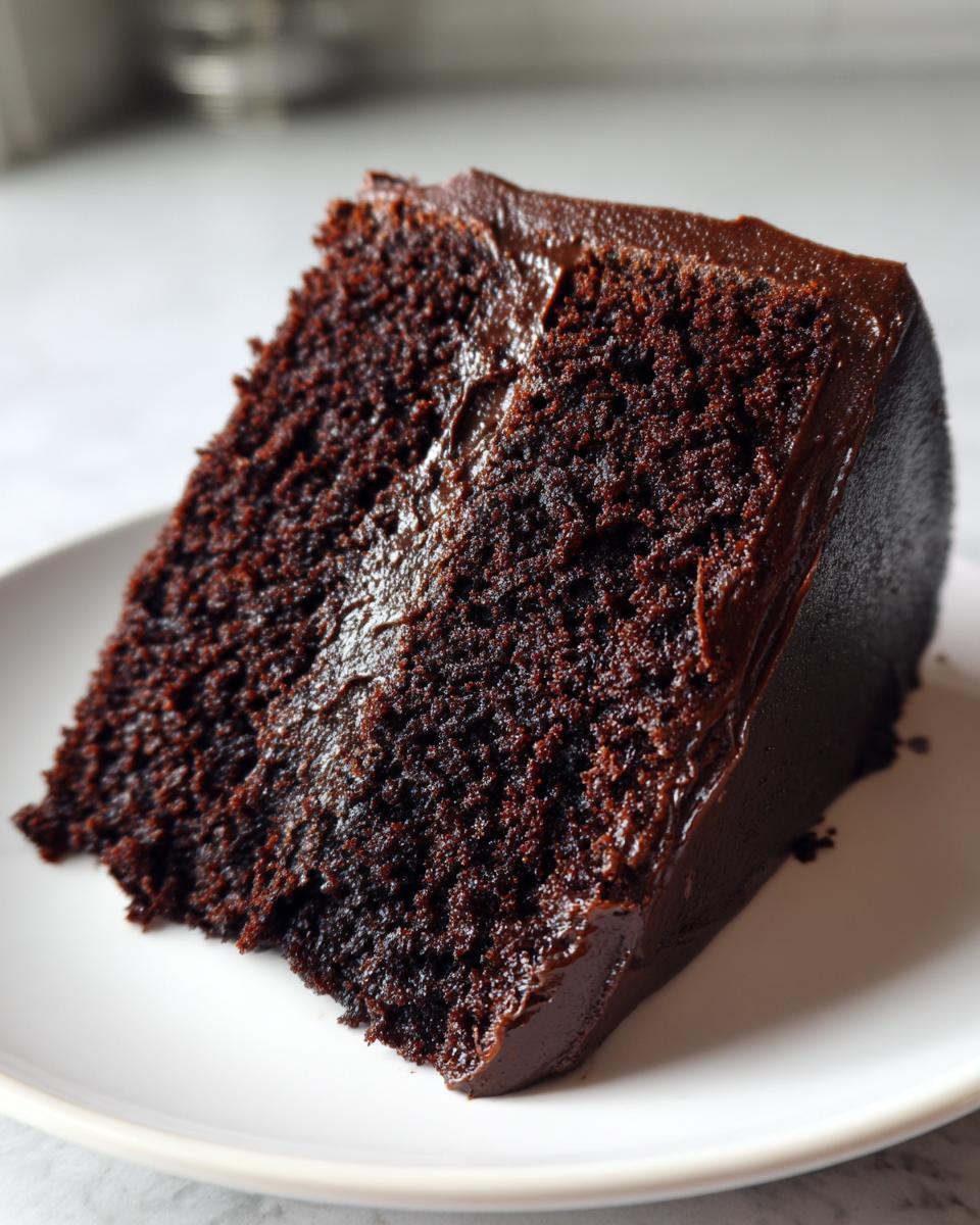 Close-up of a rich, moist slice of homemade chocolate cake covered in glossy dark chocolate frosting on a white plate.