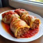 Four homemade cabbage rolls stuffed with meat and rice, served in a rich tomato sauce on a white plate.