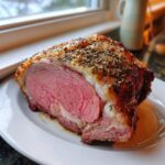 A thick slice of medium-rare prime rib recipe roast resting on a white plate with juices.