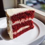A tempting slice of two-layer red velvet cake with thick white cream cheese frosting, sitting on a white plate.
