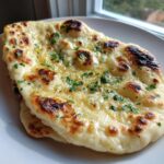 Close-up of soft, buttered naan bread, charred in spots, topped with melted butter and fresh parsley.