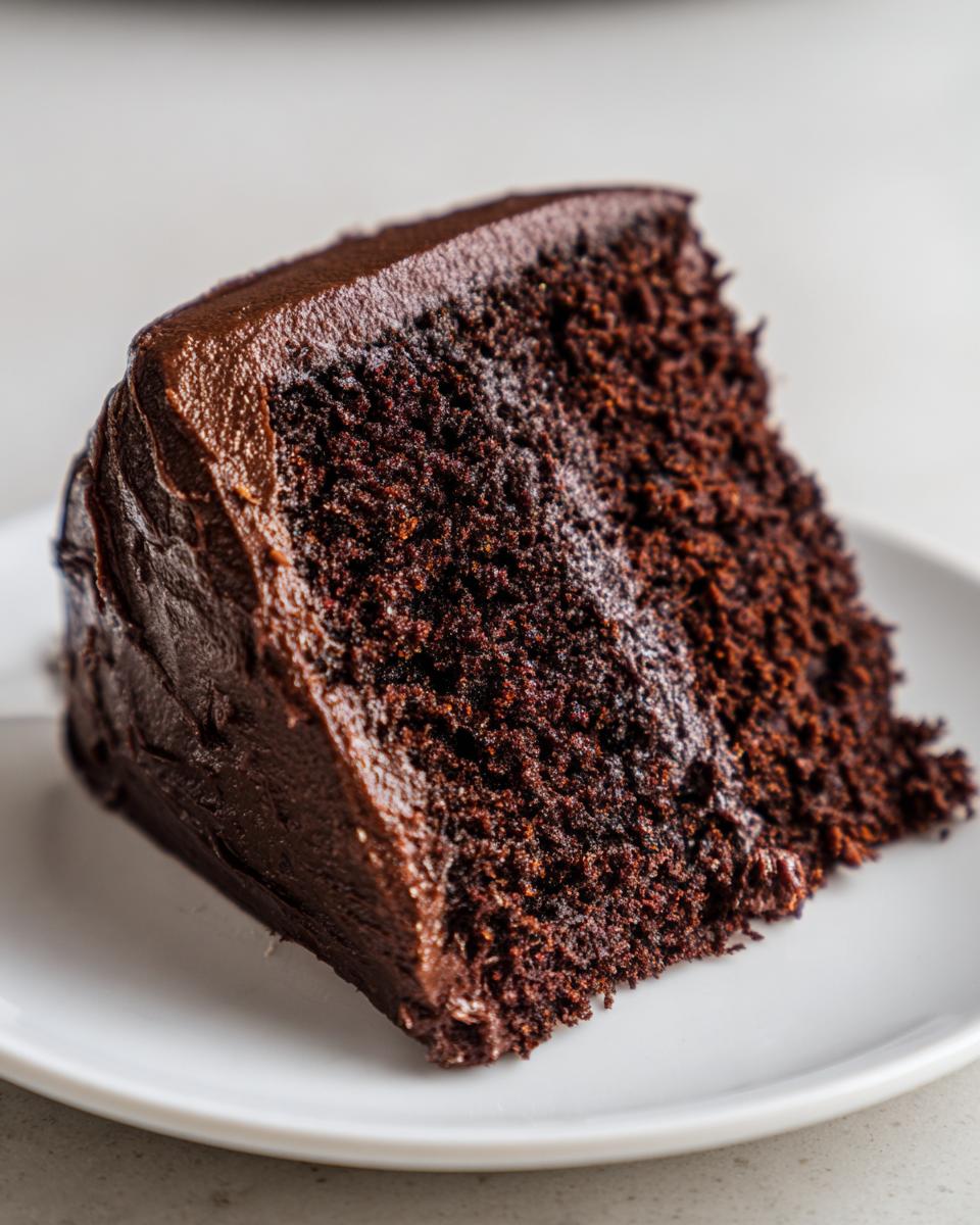 A close-up of a moist slice of homemade chocolate cake covered in thick, dark chocolate frosting on a white plate.