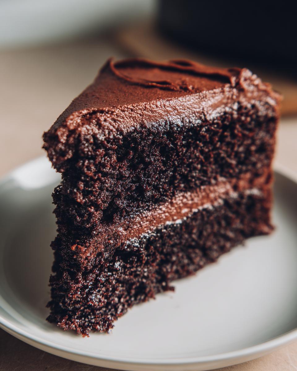 Close-up of a rich, dark slice of homemade chocolate cake layered with thick chocolate frosting.