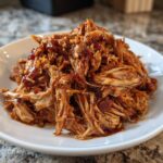 A close-up of shredded, saucy Slow Cooker Pulled Pork piled high on a white plate.