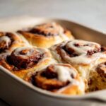 Close-up of warm, soft cinnamon buns topped with thick white icing in a baking dish.