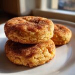 Close-up of two soft Snickerdoodle cookies stacked on a white plate, generously coated in cinnamon sugar.