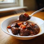 A spoonful of rich, dark crockpot beef stew with tender carrots being lifted from a white bowl.