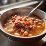 Close-up of a steaming white bowl filled with hearty navy bean soup, topped with shredded ham and diced carrots.