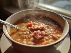 Close-up of a steaming white bowl filled with hearty navy bean soup, featuring ham chunks, carrots, and celery.
