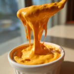 A spoonful of thick, bright orange nacho cheese sauce dripping back into a white ramekin.